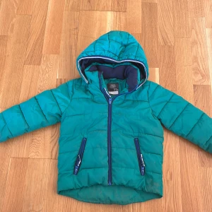Barn Turkos pufferjacka från Lindex - En barn Turkos pufferjacka från Lindex Out Wear med huva och blå detaljer vid dragkedjorna. Jackan har quiltad design, två fickor med dragkedja och ett mjukt mörkblått foder. Perfekt för kalla dagar och har en cool bergsdetalj på ryggen.
