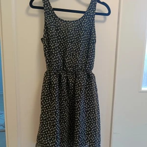 Blommig svart kortklänning med öppen rygg - Supersöt svart kortklänning med vitt blommigt mönster och en snygg öppen ryggdetalj. Klänningen har ärmlös design och markerad midja som ger en fin siluett. Perfekt för dig som gillar en lekfull och trendig stil.