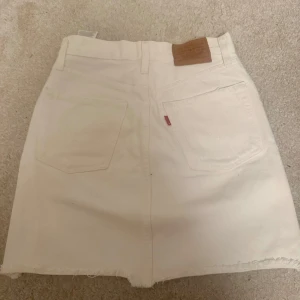 Vit jeanskjol från Levi's - Säljer en vit jeanskjol från Levi's med rå kant och slitningar framtill. Kjolen har klassiska fem fickor, knapp och dragkedja samt Levi's läderpatch bak. Använd fåtal gånger och är i fint skick. Storlek 25!
