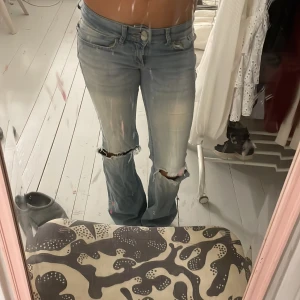 Ljusblå bootcut jeans med slitningar - Säljer ett par ljusblå jeans med bootcut passform och snygga slitningar vid knäna. Jeansen har låg midja, klassiska fem fickor och dragkedja med knapp framtill. Materialet är jeans och de har en cool, avslappnad vibe som passar perfekt till sneakers eller boots.