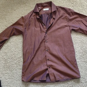 Brun skjorta med prickigt mönster från Eton - Säljer en brun långärmad skjorta från Eton med diskret prickigt mönster i lila. Skjortan har klassisk krage och knäppning framtill. Passformen är slim och materialet känns mjukt och lyxigt. Perfekt för dig som vill ha en stilren look.