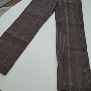 Bruna randiga linnebyxor från JOHN'S - Säljer ett par bruna linnebyxor från JOHN'S med vita vertikala ränder. Byxorna har normal passform och klassisk rak siluett. Perfekta för dig som gillar en avslappnad men stilren look. Midjemått ca. W32 (ca.82 cm) och innerbenslängd L 32 (ca. 108 cm)