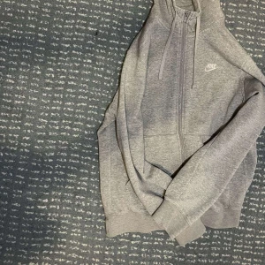 Grå hoodie från Nike med dragkedja - Säljer en klassisk grå hoodie från Nike med dragkedja framtill och två stora fickor. Tröjan har huva med snörning och mjukt fleecefoder på insidan. Perfekt för en sportig och avslappnad stil.