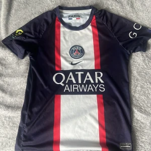 PSG Mbappé matchtröja 22/23 - Säljer en officiell Paris Saint-Germain fotbollströja med Mbappé 7 på ryggen. Tröjan är mörkblå med vita och röda detaljer, PSG-logga på bröstet och sponsortryck från Qatar Airways. Korta ärmar och tillverkad i lätt, ventilerande material. Priset kan eventuellt diskuteras.