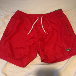 Röda badshorts från Lacoste - Snygga röda badshorts från Lacoste med vit snörning i midjan och klassisk krokodillogga. Shortsen har innerbyxa i mesh och svarta dragkedjor på fickorna. Perfekta för stranden eller poolen. I storlek S men passar mer i XS. 