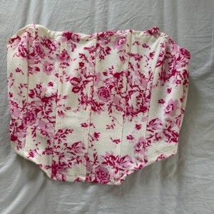 Vit och rosa blommig korsett - Supersnygg korsett med blommigt mönster i rosa och vitt. Modellen är axelbandslös och har en strukturerad form som ger en trendig siluett. Perfekt att styla till jeans eller kjol för en cool look.