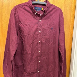Vinröd skjorta från Ralph Lauren - Säljer en vinröd skjorta från Ralph Lauren i slim fit-modell. Skjortan har långa ärmar, klassisk krage och knäppning framtill. Broderad blå logga på bröstet ger en snygg detalj. Perfekt för dig som gillar stilrena plagg med premiumkänsla.