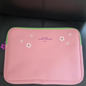 Rosa datorväska från Marc Jacobs - Säljer en söt rosa datorväska från Marc Jacobs med vita och rosa blommor på framsidan. Väskan har grön dragkedja och lila insida. Perfekt för att bära din laptop med stil. 36x26 cm