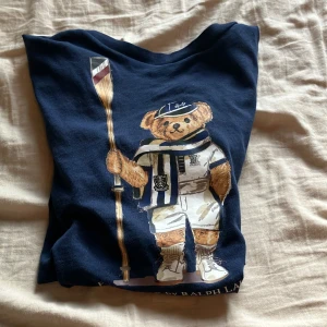 Mörkblå t-shirt med Polo Bear från Ralph Lauren - Mörkblå t-shirt från Ralph Lauren med ikoniskt Polo Bear-motiv framtill. Björnen är klädd i sportig outfit med roddtema och håller en åra. T-shirten har rund hals och korta ärmar. Perfekt för dig som gillar klassisk och lekfull stil.