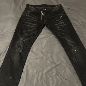 Svarta jeansbyxor från Dsquared2 - Säljer ett par svarta jeansbyxor från Dsquared2 med slitna detaljer och små hål på benen. Byxorna har dragkedja framtill och klassisk femficksdesign. Märkeslapp bak i midjan och snygg, modern look.