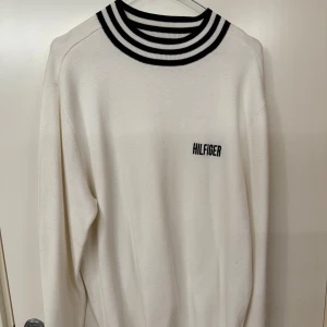 Vit långärmad tröja från Tommy Hilfiger - Snygg vit långärmad tröja från Tommy Hilfiger med svart broderad logga på bröstet. Tröjan har en unik ribbad krage med svarta och vita ränder som ger en sportig känsla. Perfekt för en stilren och avslappnad look.