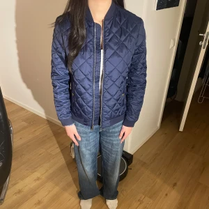 Mörkblå quiltad jacka från Polo Ralph Lauren - Snygg mörkblå quiltad jacka från Polo Ralph Lauren med dragkedja framtill och ribbade muddar. Jackan har en klassisk bomberjacke-look och guldiga knappar vid fickorna. Perfekt för dig som vill ha en stilren och tidlös jacka. Modellen på bilden är 164, skick 8/10