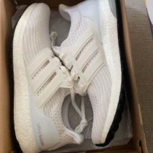 Vita Adidas Ultraboost sneakers - Säljer ett par fräscha vita Adidas Ultraboost sneakers med stickad ovandel och klassiska tre ränder på sidan. Skorna har snörning och en bekväm, responsiv sula som ger extra studs i steget. Perfekta för både träning och vardag.