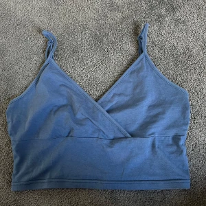 Blå omlott linnetopp - Säljer en blå omlott linnetopp med smala axelband som knyts upptill. Toppen har en enkel och stilren design med v-ringning och är tillverkad i mjukt bomullsmaterial. Perfekt för varma dagar eller som lager under andra plagg.
