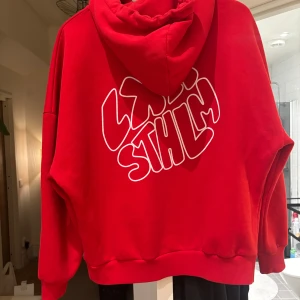 Röd hoodie med tryck på ryggen - Säljer en röd hoodie med stor vit text på ryggen. Tröjan har huva och en avslappnad passform, perfekt för en streetwear-look.  Från lxa