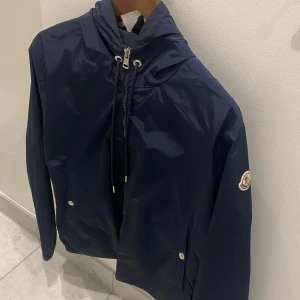 Moncler windbreaker - Säljer en snygg mörkblå vindjacka från Moncler. Jackan har en dragkedja framtill och en justerbar huva med röd och vit randdetalj. Den är långärmad och har Monclers logga på ärmen. Väldigt fint skick 🤝 Storlek 2 motsvarar M 