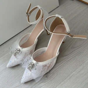 Vita spetsiga pumps med pärldetaljer - Säljer ett par vita pumps med spetsig tå, klack och vacker spets över hela skon. Skorna har en dekorativ rosett framtill med pärlor och strass, samt justerbart ankelband. Perfekta för bal eller student! Klackhöjd 6cm