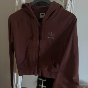 Brun hoodie med dragkedja från Adidas - Säljer en brun hoodie från Adidas med dragkedja och huva. Tröjan har långa ärmar, två fickor framtill och Adidas-logga i strass på bröstet. Perfekt för en avslappnad stil och enkel att matcha med jeans eller träningsbyxor.
