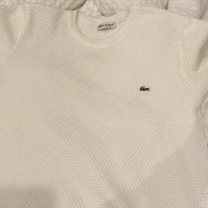 Vit stickad tröja från Lacoste - Säljer en klassisk vit stickad tröja från Lacoste med rund halsringning och det ikoniska gröna krokodilmärket broderat på bröstet. Perfekt för en stilren och enkel look.