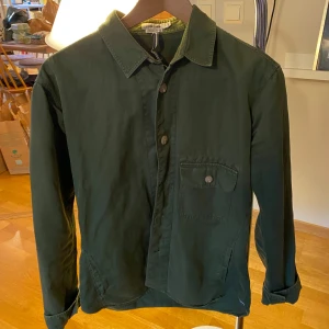 Grön Overshirt från J.Lindeberg - Säljer en grön skjorta från J.Lindeberg i regular fit. Skjortan har klassisk krage, bröstficka med knapp och knäppning framtill. Perfekt för dig som gillar stilrena plagg med enkel design.