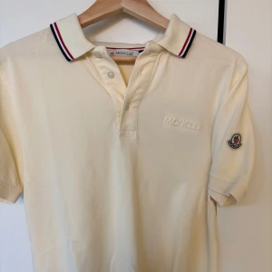 Moncler piké - Snygg vit piké t-shirt från Moncler med broderad logga på bröstet och ärmen. Kragen har stilrena ränder i rött och blått för en sportig look. Perfekt till jeans eller shorts.