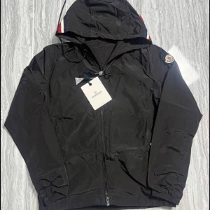 Svart vindjacka från Moncler - Svart vindjacka från Moncler med huva och dragkedja framtill. Jackan har Moncler-logga på ärmen och detaljer i rött, vitt och blått på huvan. Perfekt för blåsiga dagar och enkel att matcha med det mesta.