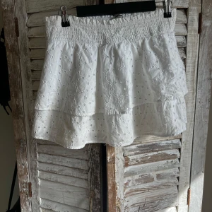 Vit volangkjol från Gina Tricot - Supersöt vit kjol från Gina Tricot med broderade detaljer och volanger. Kjolen har smock i midjan och är tillverkad i bomull, perfekt för en fräsch och somrig look. 
