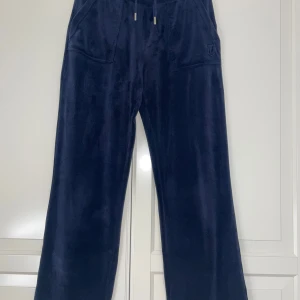Mörkblå mjukisbyxor från Juicy Couture - Mörkblå mjukisbyxor från Juicy Couture med breda ben och dragsko i midjan. Byxorna har fickor fram och bak samt broderad logga på ena fickan. Mjukt och glansigt material som känns lyxigt och bekvämt. Litet hål vid vänstra benet!