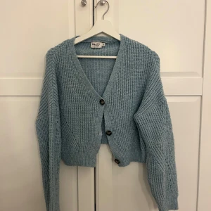 Ljusblå stickad kofta från NA-KD - Supermysig ljusblå stickad kofta från NA-KD med v-ringning och stora knappar framtill. Koftan är croppad och har långa ärmar. Perfekt för lager-på-lager och en chill look.