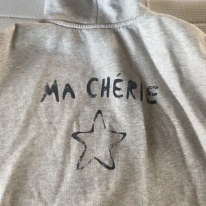 Grå hoodie med tryck 'Ma Chérie' och stjärna - Säljer en grå hoodie med svart tryck där det står 'Ma Chérie' och en stjärna på bröstet. Tröjan har huva med dragsko och långa ärmar.   Ska tvättas innan om jag får sålt den jag använde den lite förr men aldrig lägre