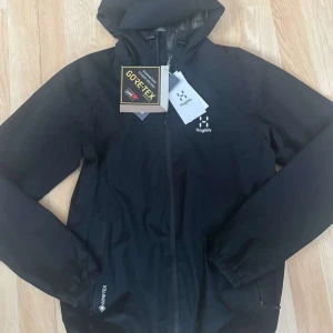 Svart skaljacka från Haglöfs med huva - Svart skaljacka från Haglöfs med huva och dragkedja framtill. Jackan är tillverkad i GORE-TEX-material och har diskret logga på bröstet. Perfekt för utomhusaktiviteter och skyddar mot regn och vind.