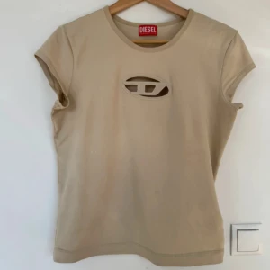 Beige t-shirt med cut-out från Diesel - Säljer en beige t-shirt från Diesel med unik cut-out detalj och broderad logga framtill. T-shirten har rund halsringning och korta ärmar. Perfekt för dig som vill sticka ut med en cool och trendig look.