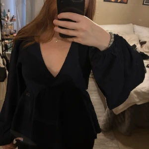 Mörkblå blus med v-ringning  - Säljer en mörkblå blus med djup v-ringning och långa, vida ärmar. Blusen har en lätt volang nedtill och är perfekt för dig som gillar en luftig och stilren look.