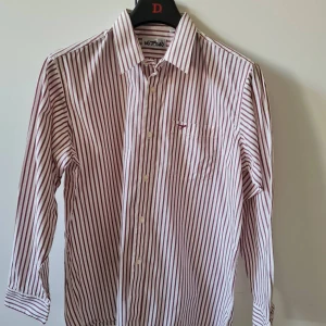 Randig skjorta från Hubao strl 41 (L) - Säljer en klassisk skjorta från Hubao med vita och vinröda/bruna ränder. Skjortan har lång ärm, knappar framtill och en bröstficka med liten broderad detalj. Material: bomull. Bröstmått 114cm. Storlek 41 (L). Kan hämtas i Vallentuna.