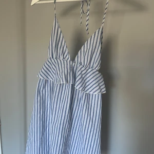 Blåvit randig klänning med volang - Säljer en blåvit randig klänning med smala axelband som knyts i nacken och djup v-ringning. Klänningen har volangdetalj under bysten och är tillverkad i ett lätt material, perfekt för varma dagar.