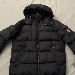 Svart dunjacka från Moncler med huva - Säljer en svart dunjacka från Moncler köpt från merchsweden (kvitto finns) denna vinter med huva och dragkedja. Jackan har två snedställda fickor med dragkedja framtill och en ficka med knapp på ärmen där Moncler-loggan sitter. Klassisk pufferstil med quiltade sektioner. Endast använda fåtal gånger pga storlek lite stor. 