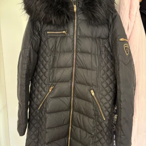 Svart lång dunjacka med fuskpäls från Rockandblue - Säljer en svart lång dunjacka från Rockandblue med fuskpäls runt kragen. Jackan har guldiga dragkedjor, tre fickor framtill och ett Rockandblue-märke på ärmen. Quiltad design ger en snygg och modern look. Jackan är i sttorlek 42 men passar även som M.