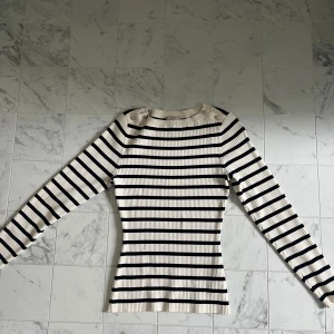 Randig ribbad långärmad tröja - Säljer en vit och svart ribbad långärmad tröja med smal passform. Tröjan har rund halsringning och dekorativa knappar på axeln. Perfekt för dig som gillar klassiska ränder och en figurnära stil.