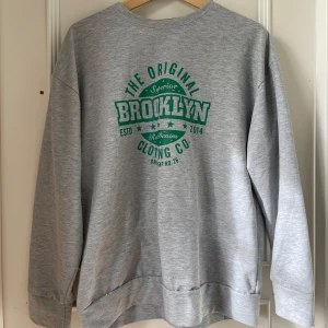 Grå sweatshirt med tryck från Not Your Honey - Grå sweatshirt med grönt tryck där det står 'Brooklyn'. Tröjan har rund halsringning och långa ärmar. Perfekt för en avslappnad stil och enkel att matcha med jeans eller joggers.