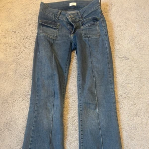 Blå jeans med utsvängda ben från Nelly - Säljer ett par blå jeans från Nelly med utsvängda ben och stora fickor framtill. Jeansen har låg midja och klassisk dragkedja med dubbla knappar. Perfekta för dig som gillar retrostil och vill ha något unikt i garderoben. Ganska slitna vid slutet av benen