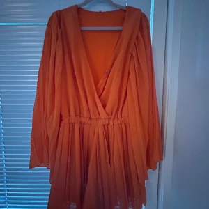 Orange omlottklänning med vida ärmar - Säljer en kort orange omlottjumpsuit med v-ringning och långa, vida ärmar. Klänningen har resår i midjan och ett plisserat, luftigt tyg som ger en flowy känsla. Perfekt för dig som vill sticka ut med färg!