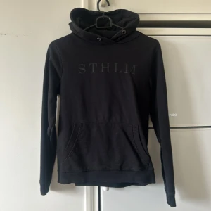 Svart hoodie med STHLM-tryck - Svart hoodie med trycket 'STHLM' på bröstet. Tröjan har huva med dragsko och en stor magficka framtill. Långa ärmar och mjukt material gör den perfekt för kyligare dagar.