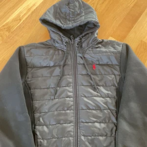 Svart cardigan från Polo Ralph Lauren - Säljer en svart hoodie från Polo Ralph Lauren med quiltad framsida och ärmar i mjukare material. Jackan har huva, dragkedja och en liten röd logga på bröstet. Perfekt för dig som gillar sportig och stilren look.