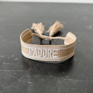 Champange färgat dior armband - Snyggt beige armband med broderad vit text 'J'ADORE' och dekorativa tofsar i ändarna. Justerbar passform med knytband. Perfekt accessoar för att lyfta din stil.