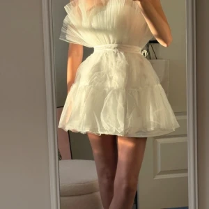 Vit tyllklänning med volang - Supersöt vit kortklänning i tyll med volangdetaljer och markerad midja. Klänningen är ärmlös och har en fluffig, luftig känsla som ger en riktigt drömmig look. Perfekt för dig som vill sticka ut med en romantisk stil.