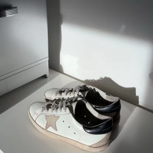 Golden Goose skor - Golden Goose sneakers i vitt skinn med beige stjärna på sidan och svarta hälpartier. Skorna har snörning och platt sula. Klassisk och stilren modell med en unik vintage-look och diskreta detaljer.