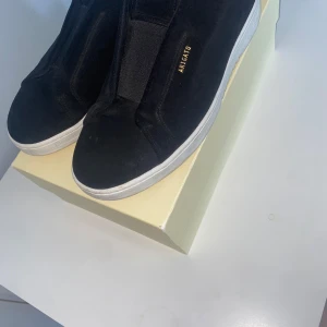 Svarta sneakers från Axel Arigato - Säljer ett par svarta sneakers från Axel Arigato med vit sula och diskret guldfärgad logga på sidan. Skorna har en stilren design utan snörning och är tillverkade i mocka. Perfekta för dig som gillar minimalistisk stil.