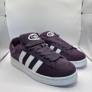 Lila Adidas Campus 00s sneakers - Säljer ett par lila Adidas Campus 00s sneakers med klassiska vita ränder på sidorna och vit logga på plösen och hälen. Skorna har en ovandel i mocka och vit platt sula. Perfekta för dig som gillar retrostil och streetwear. Aldrig använda, helt nya! Lite små i storleken.