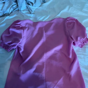 Rosa kortärmad blus med puffärm - Säljer en söt rosa blus med v-ringning och korta puffärmar med resår. Blusen har en lätt struktur i tyget och en feminin siluett. Perfekt för dig som gillar färg och vill sticka ut lite extra.