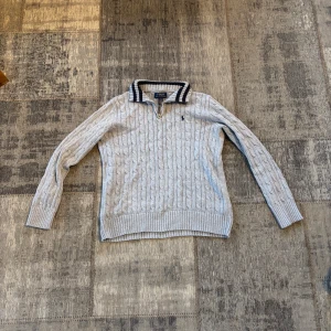 Ljusgrå kabelstickad tröja från Polo Ralph Lauren - Kabelstickad ljusgrå tröja från Polo Ralph Lauren med klassisk krage och dragkedja vid halsen. Tröjan har långa ärmar och en liten broderad logga på bröstet. Perfekt för en stilren och avslappnad look.